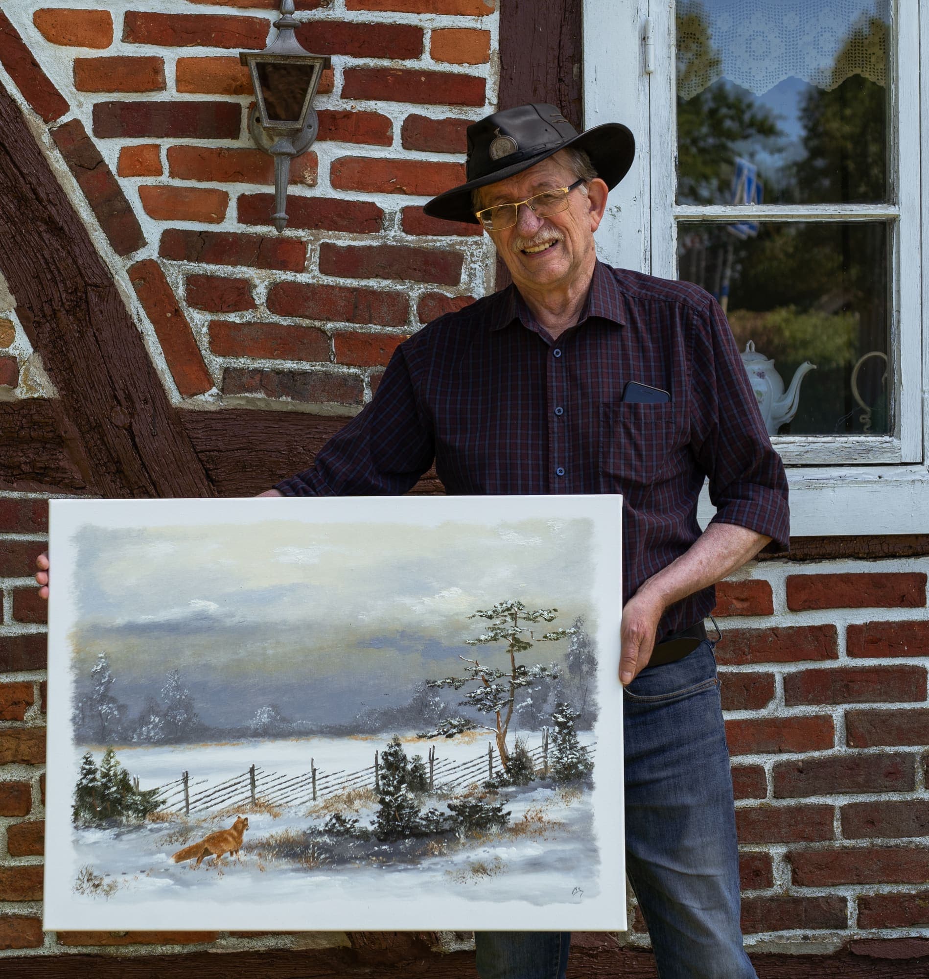 Konstnären Ingvar Stendahl står framför sitt korsvirkeshus på Skomakaregården och håller upp en vintermålning med en räv