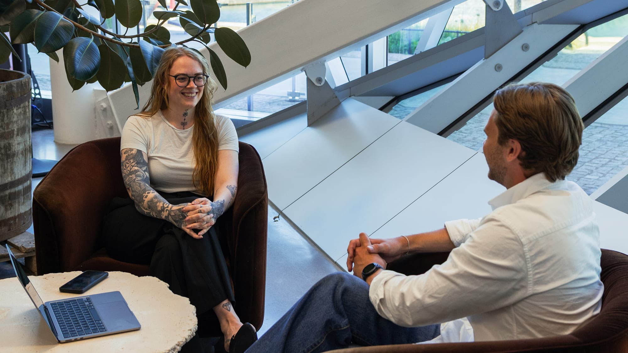 Mikaela Rasmusson och Theo Skogsberg sitter i varsin fåtölj och samtalar över ett runt bord med en uppfälld laptop, i en ljus lokal med stora fönster mot vattnet