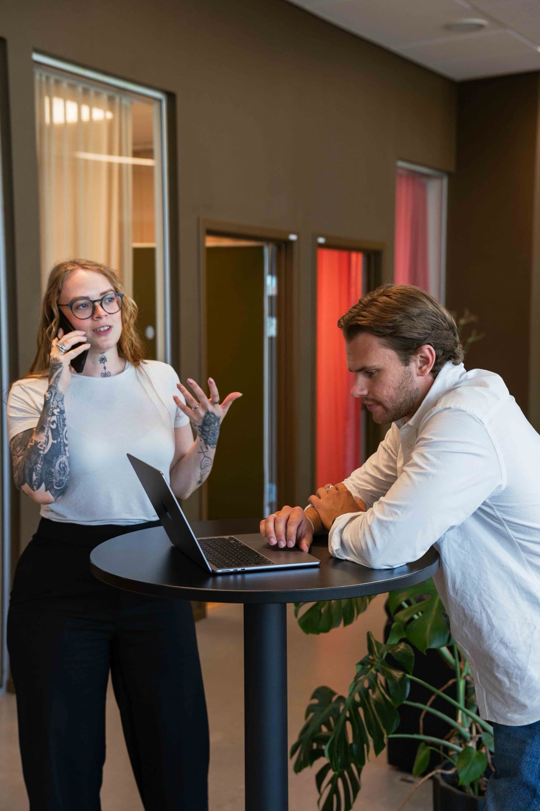 Mikaela står vid ett ståbord och talar i mobiltelefon medan Theo lutar sig mot bordet och tittar på en öppen laptop.