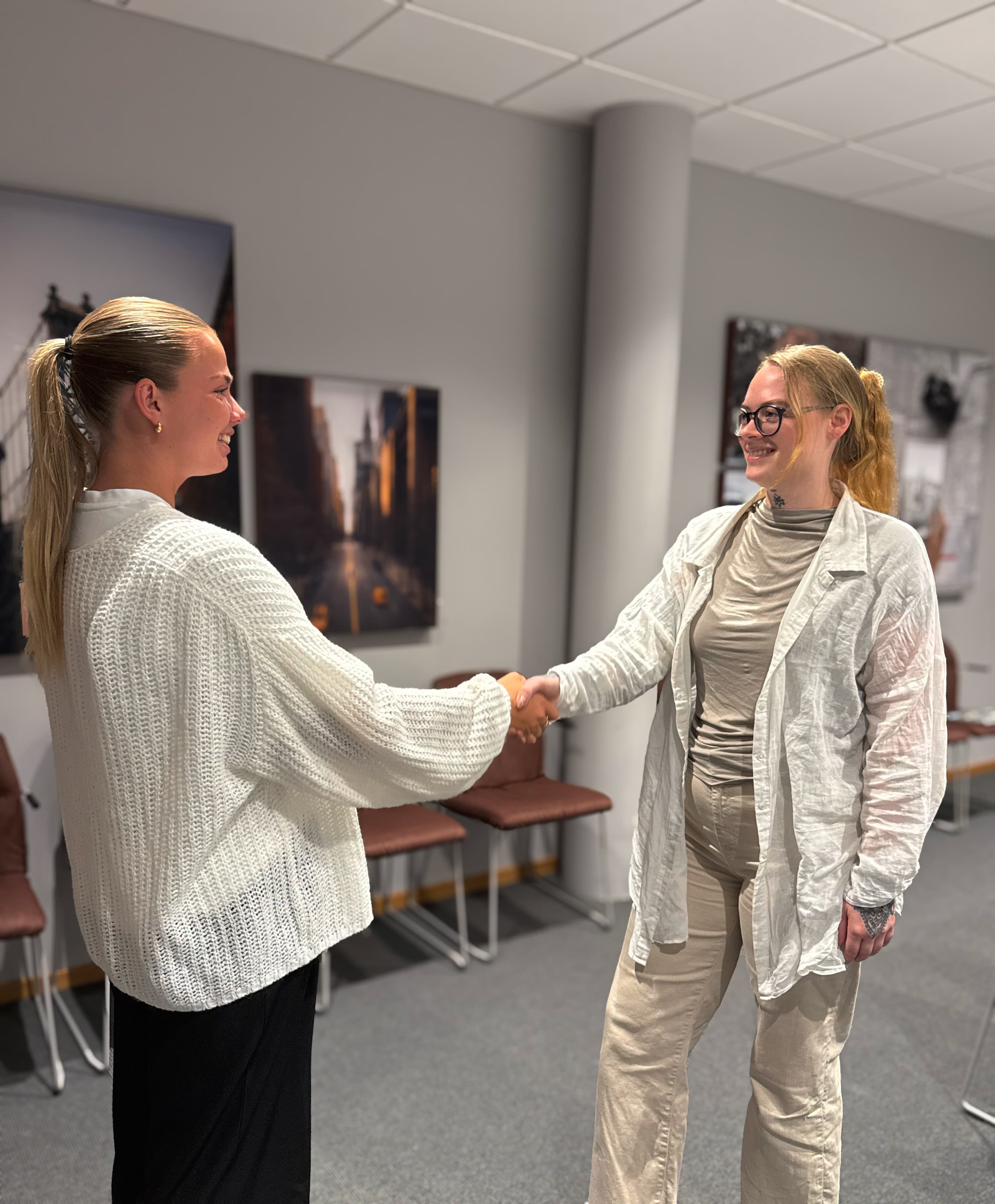 Mikaela Rasmusson skakar hand med en kund i ett ljust mötesrum med inramade stadsfotografier på väggarna. Båda ler.