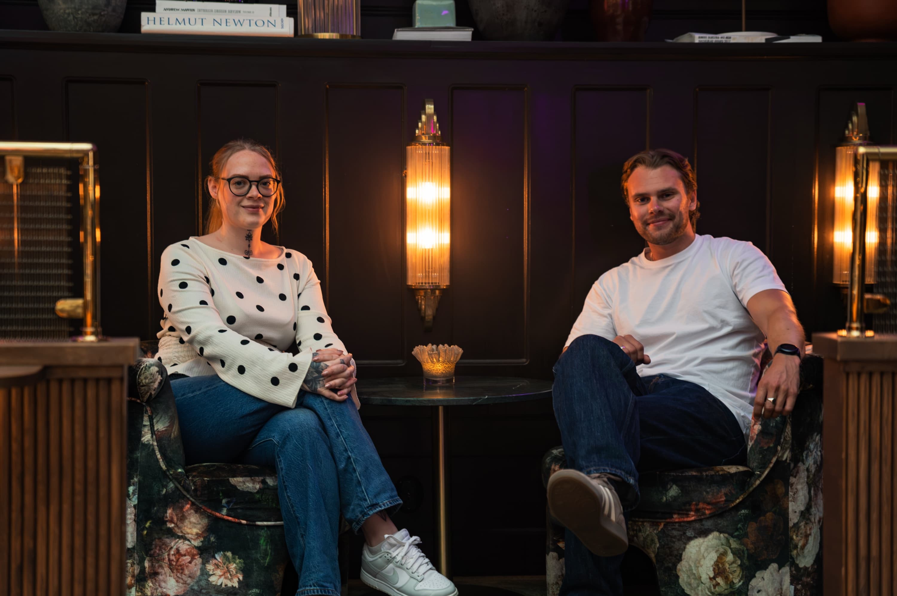 Theo och Mikaela sitter i var sin fåtölj i en mörkt inredd hotellounge med varm belysning.